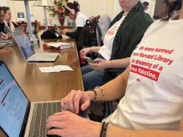 Student protestors wearing white shirts with red text stating "We were banned from Harvard Library for dreaming of a Free Palestine" working on their laptops.