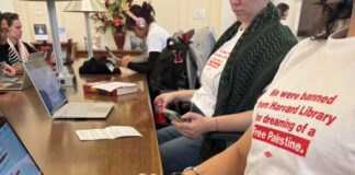 Student protestors wearing white shirts with red text stating "We were banned from Harvard Library for dreaming of a Free Palestine" working on their laptops.