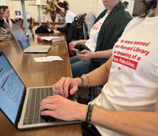 Student protestors wearing white shirts with red text stating "We were banned from Harvard Library for dreaming of a Free Palestine" working on their laptops.