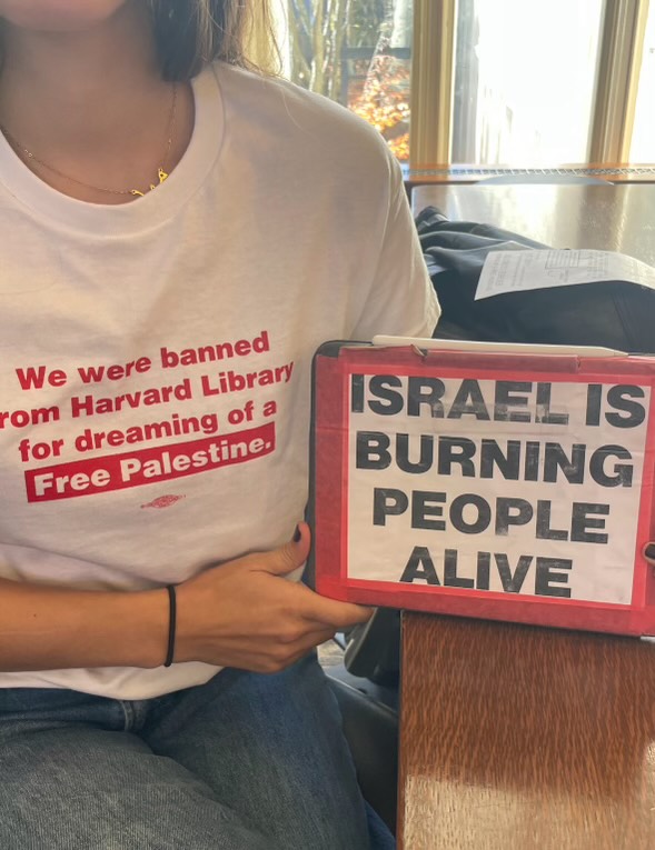 A student protestor wearing a white shirt with red text stating "We were banned from Harvard Library for dreaming of a Free Palestine" and displaying a message on their tablet in black text on white background stating "ISRAEL IS BURNING PEOPLE ALIVE" in all caps.