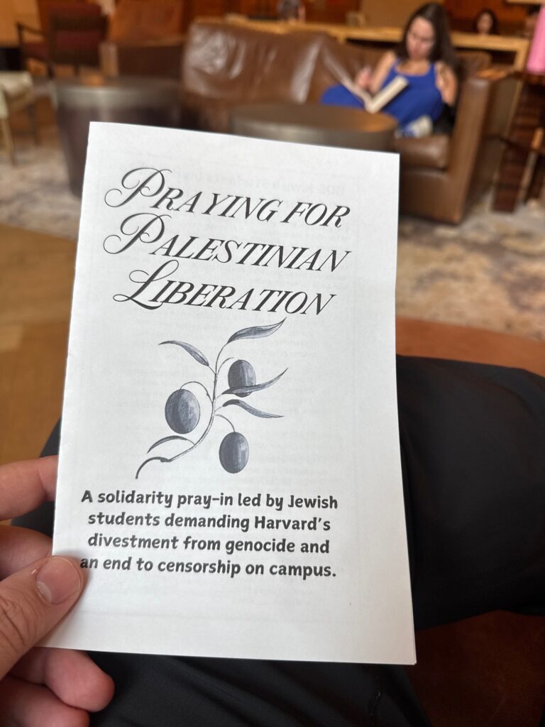 A white prayer book in a student's hand stating "Praying for Palestinian Liberation" in italicized text, followed by a caption "A solidarity pray-in led by Jewish students demanding Harvard's divestment from genocide and an end to censorship on campus."