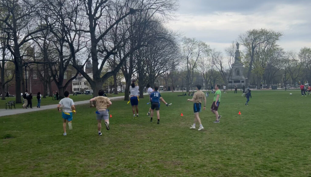 Several flag football players running in Cambridge Common park in the midst of a match. Adi Kumar, HLS '26, reaches his arms out for a catch in the endzone.